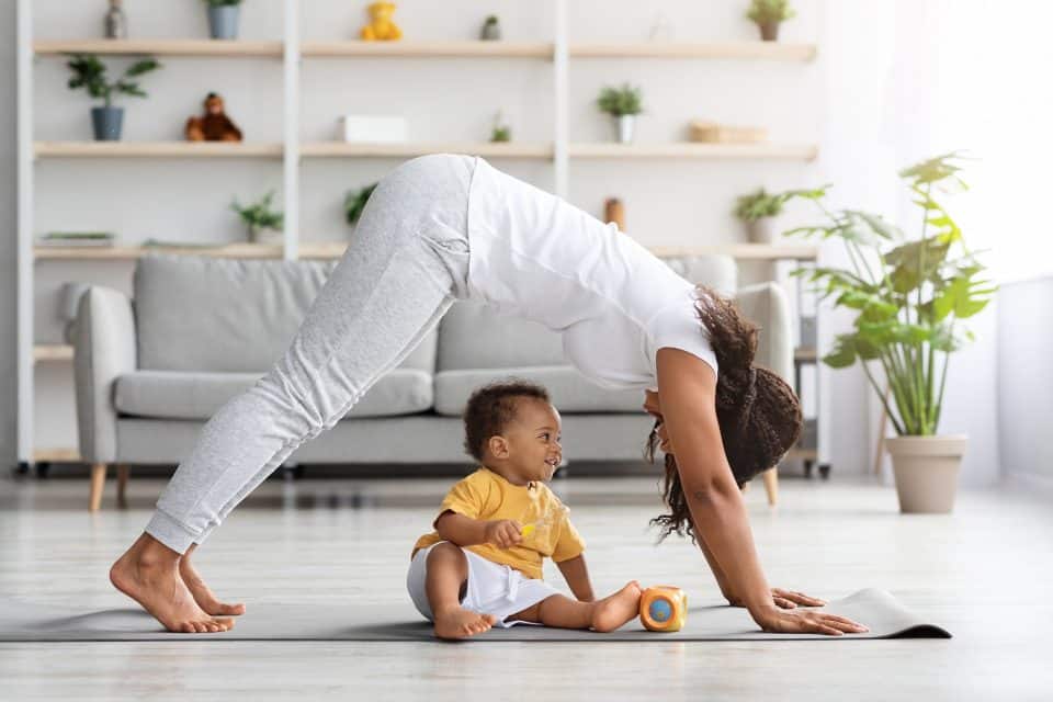 A Black mother wearing a gray shirt and pants doing a downward dog yoga pose over her infant son, who is wearing a yellow shirt and white shorts.
