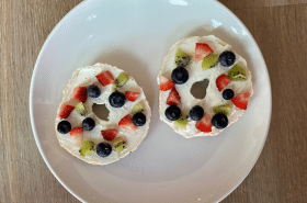 two bagels with cream cheese topped with strawberries, blueberries, and kiwi to make bagel fruit pizza