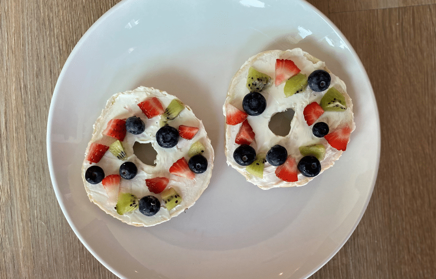 two bagels with cream cheese topped with strawberries, blueberries, and kiwi to make bagel fruit pizza