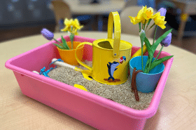 garden sensory bin for kids with brown rice, small watering can, fake flowers, rubber worms, and kid-friendly gardening tools