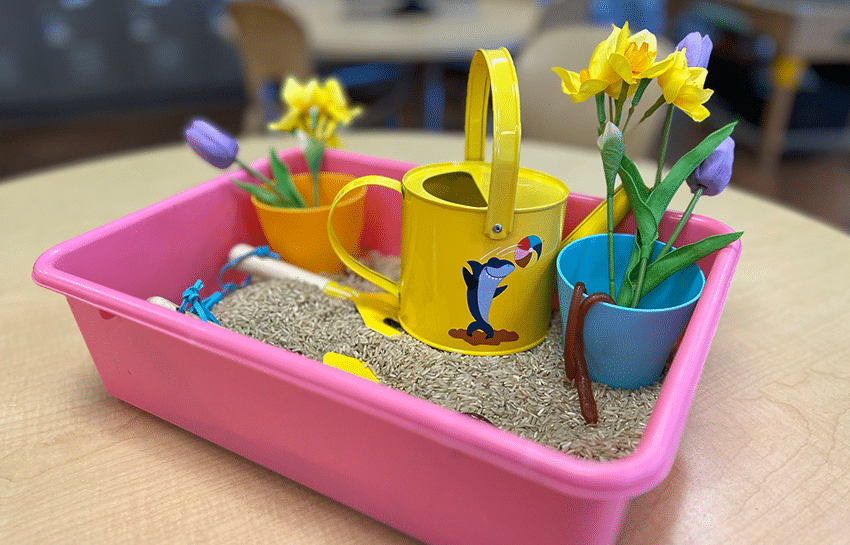 garden sensory bin for kids with brown rice, small watering can, fake flowers, rubber worms, and kid-friendly gardening tools