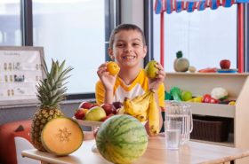 Preschool child holding up healthy fruits while eating at school